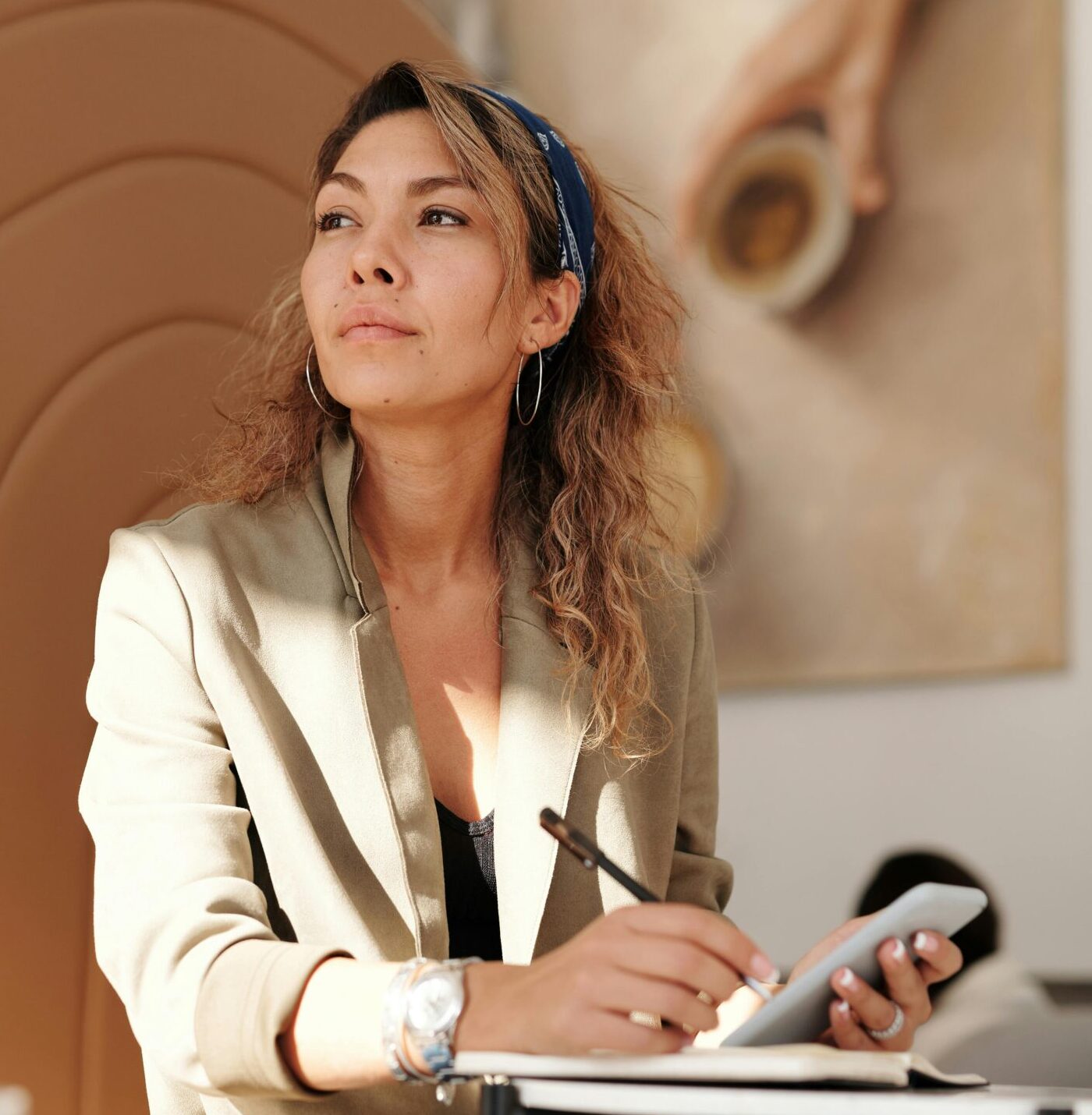 Confident woman in blazer taking notes with pen and smartphone in cafe.